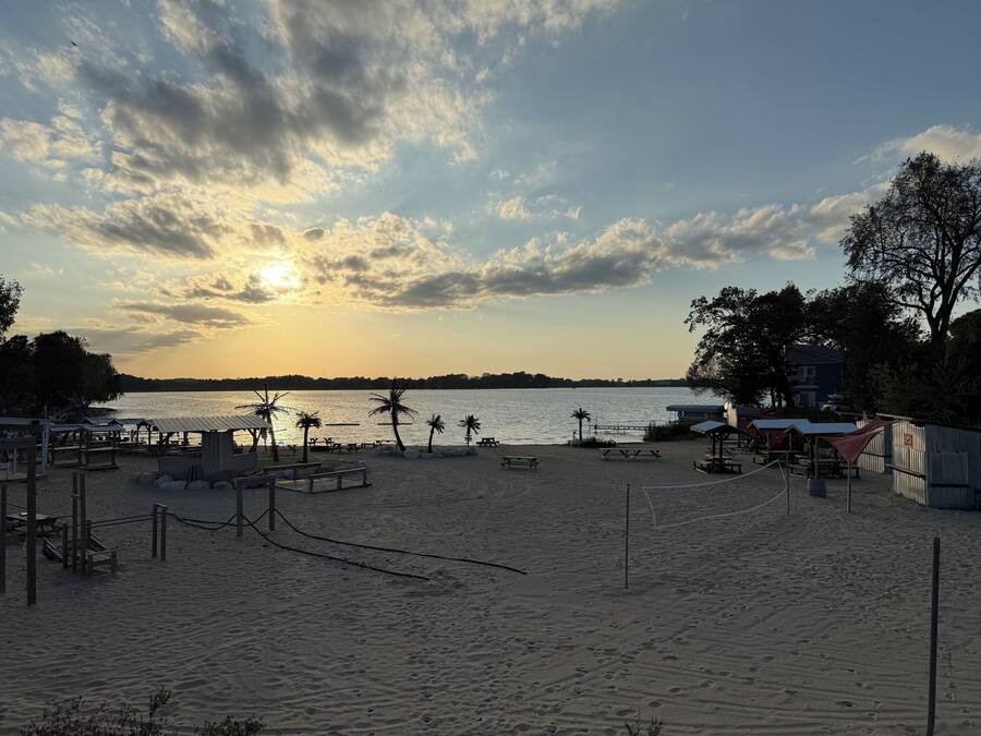Una playa con mesas de picnic y sombrillas, un muelle y un edificio al fondo.
