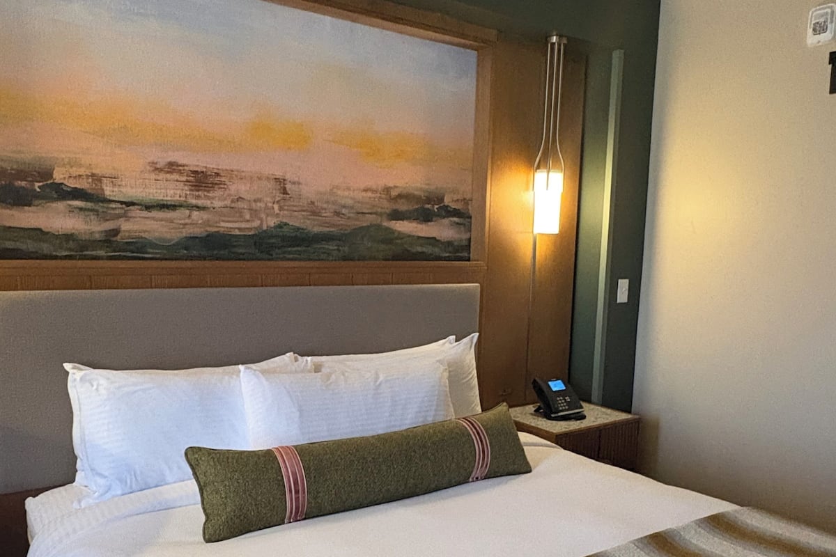 A hotel room with a large bed, a painting on the wall, a nightstand with a lamp, and a telephone.