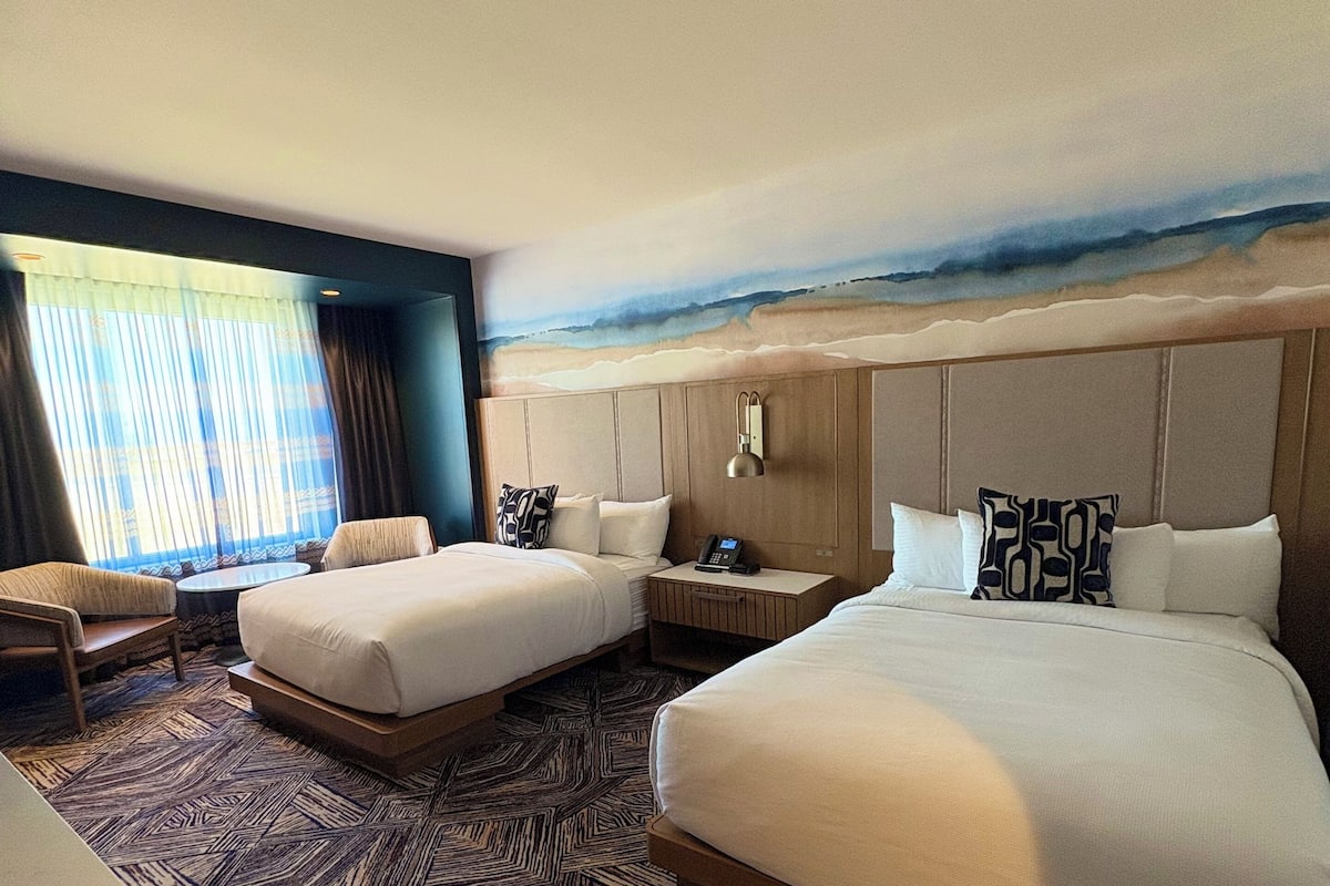A hotel room with two beds, a sofa, a desk, and a large window with a view of the beach.