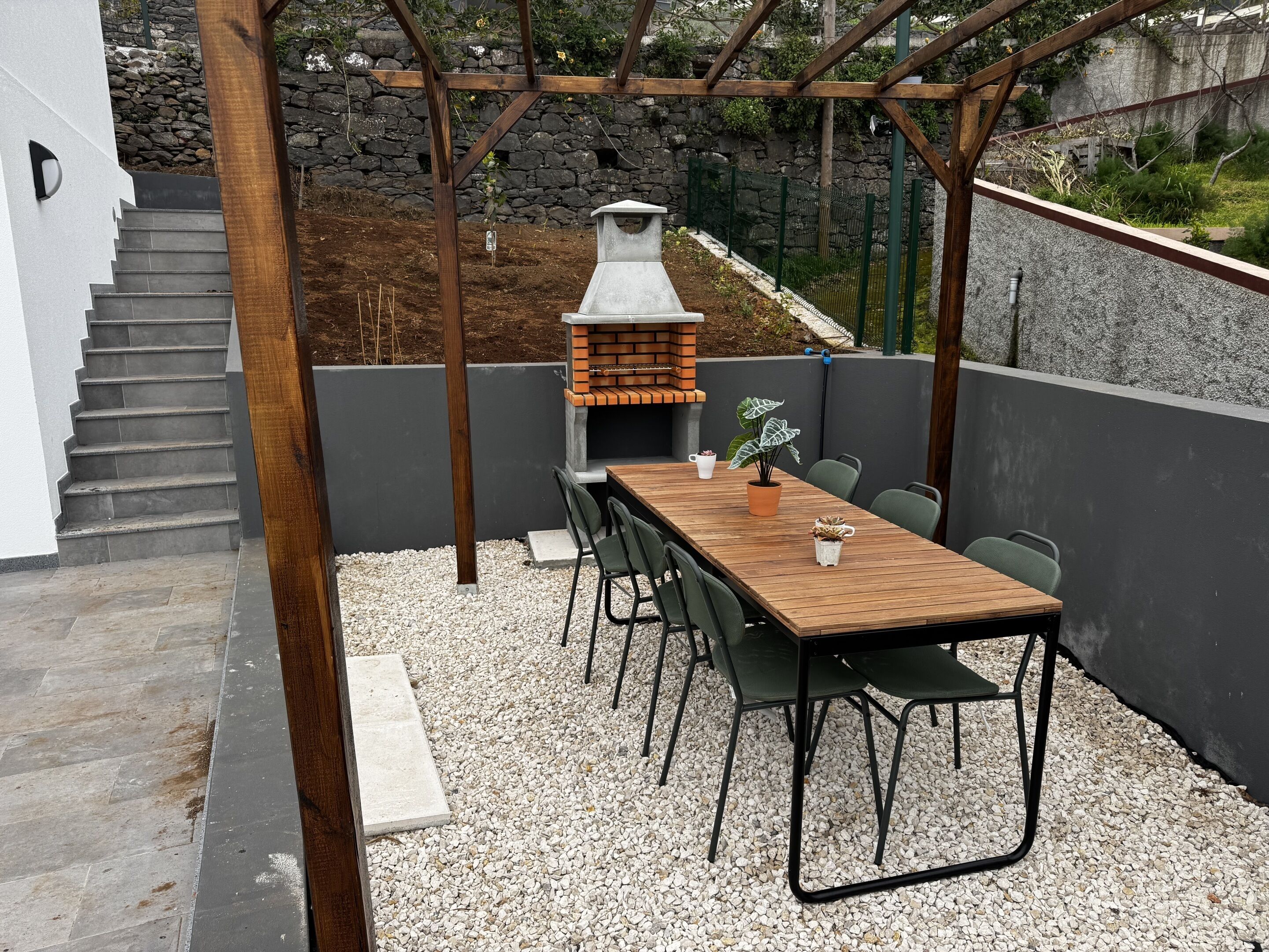 A patio with a wooden table and chairs, a brick barbecue, and a stone wall.