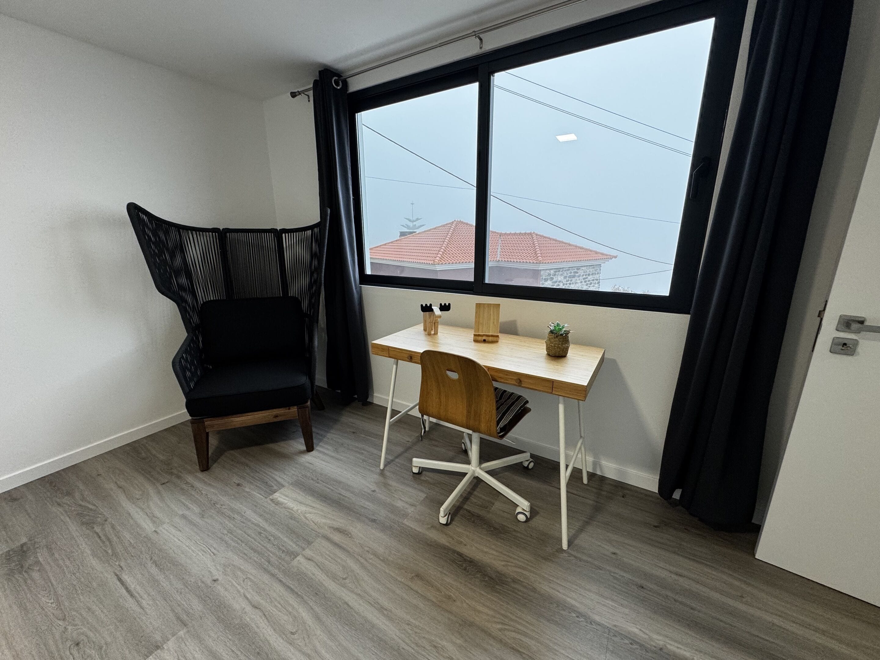 A room with a desk, a chair, a window with curtains, and a view of buildings outside.