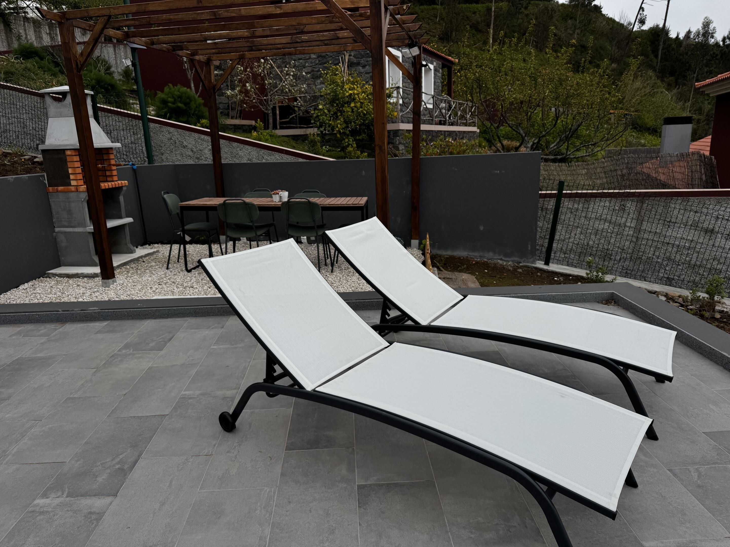 Deux fauteuils de jardin avec des coussins blancs, une pergola, une cheminée en pierre et un coin repas avec des chaises et une table.