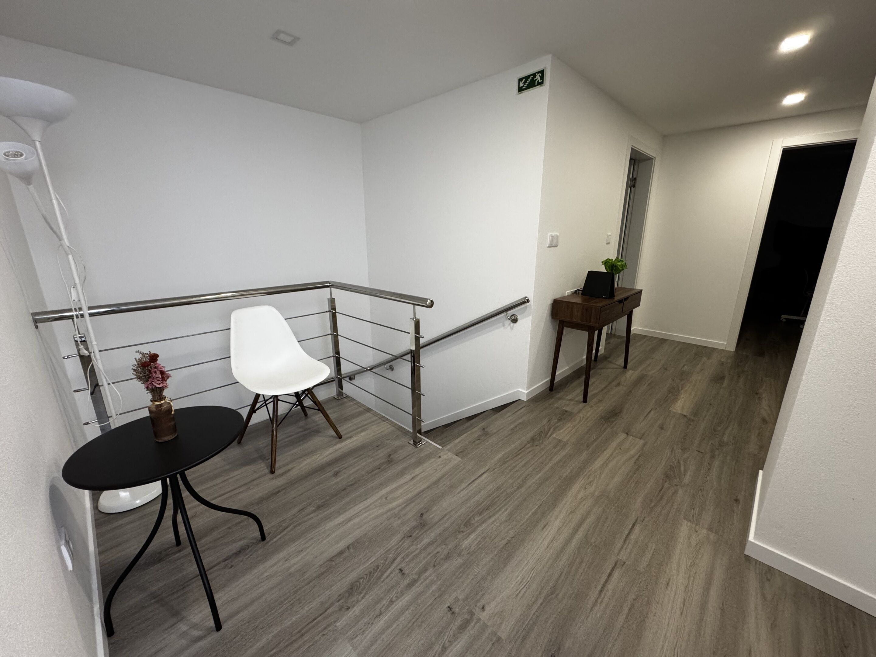 Un pasillo con pared blanca, barandilla metálica, una silla blanca, una mesita con un florero y un banco de madera.