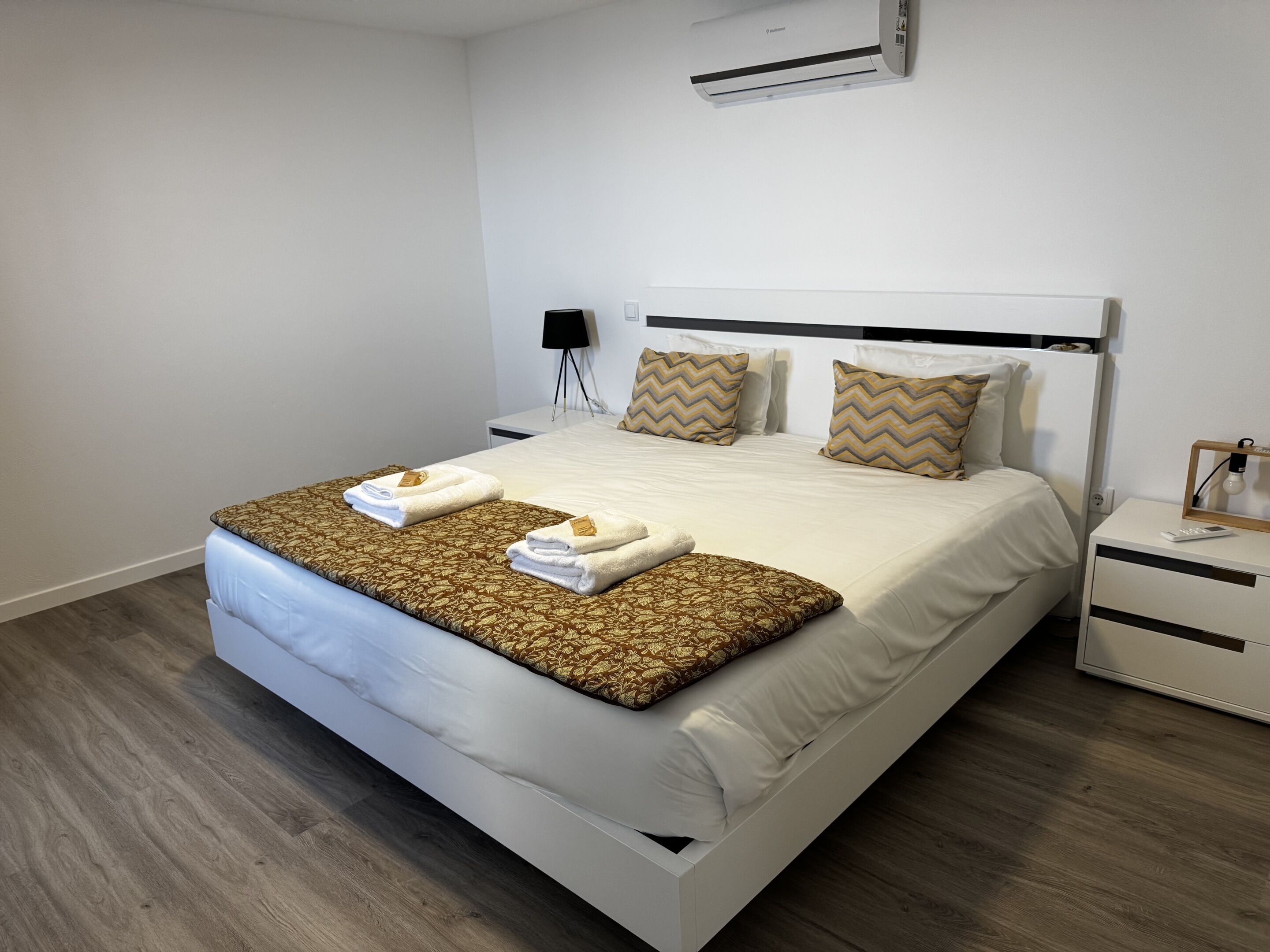A neatly made bed with pillows, a bedside table with a lamp, and a wall-mounted air conditioner.