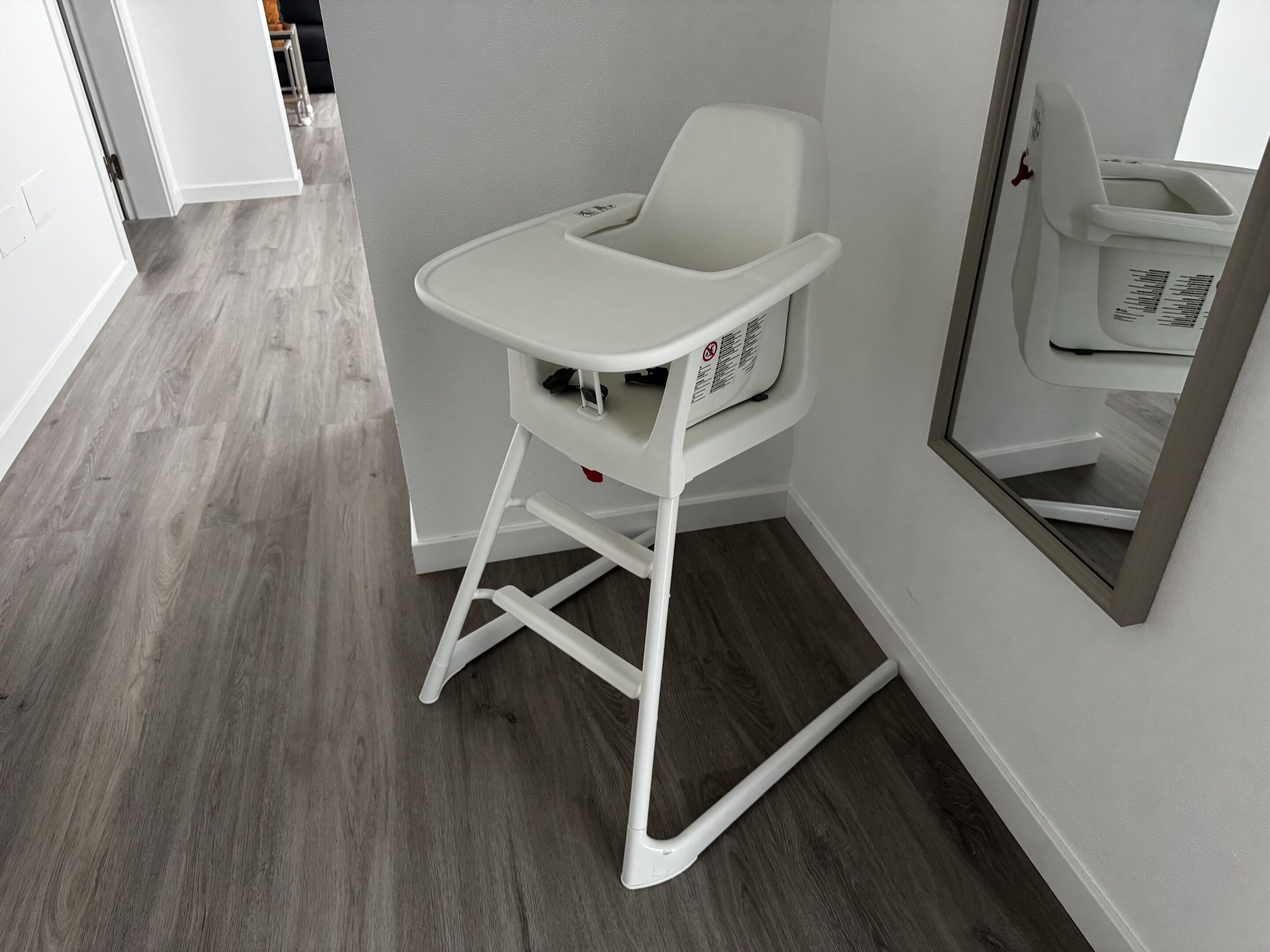 A white high chair with a tray table against a wall, a mirror reflecting a white toilet, and a wooden floor.