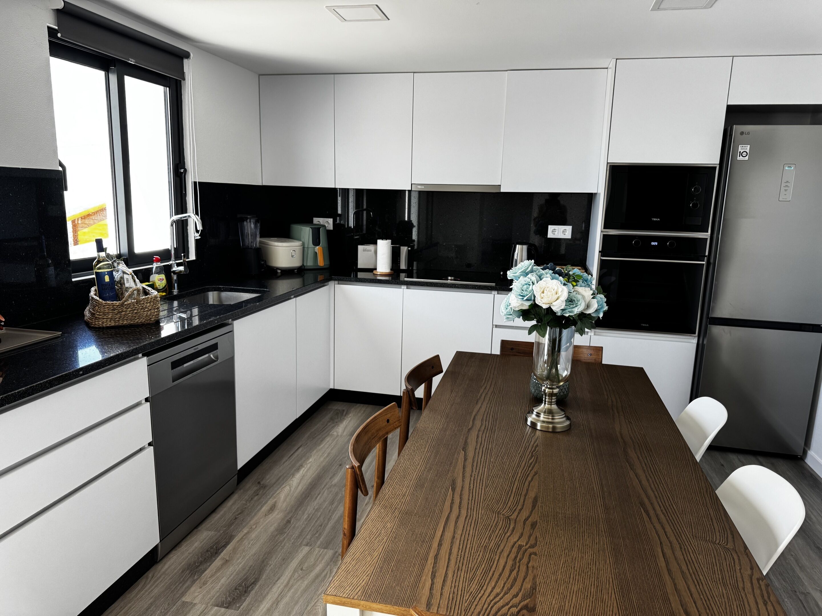 A modern kitchen with white cabinets, a wooden dining table, and a vase of flowers.