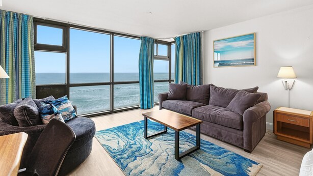 A modern living room with a large window, a sofa, a coffee table, and a lamp. - Oceanside Inn 313 (Daytona Beach Shores)