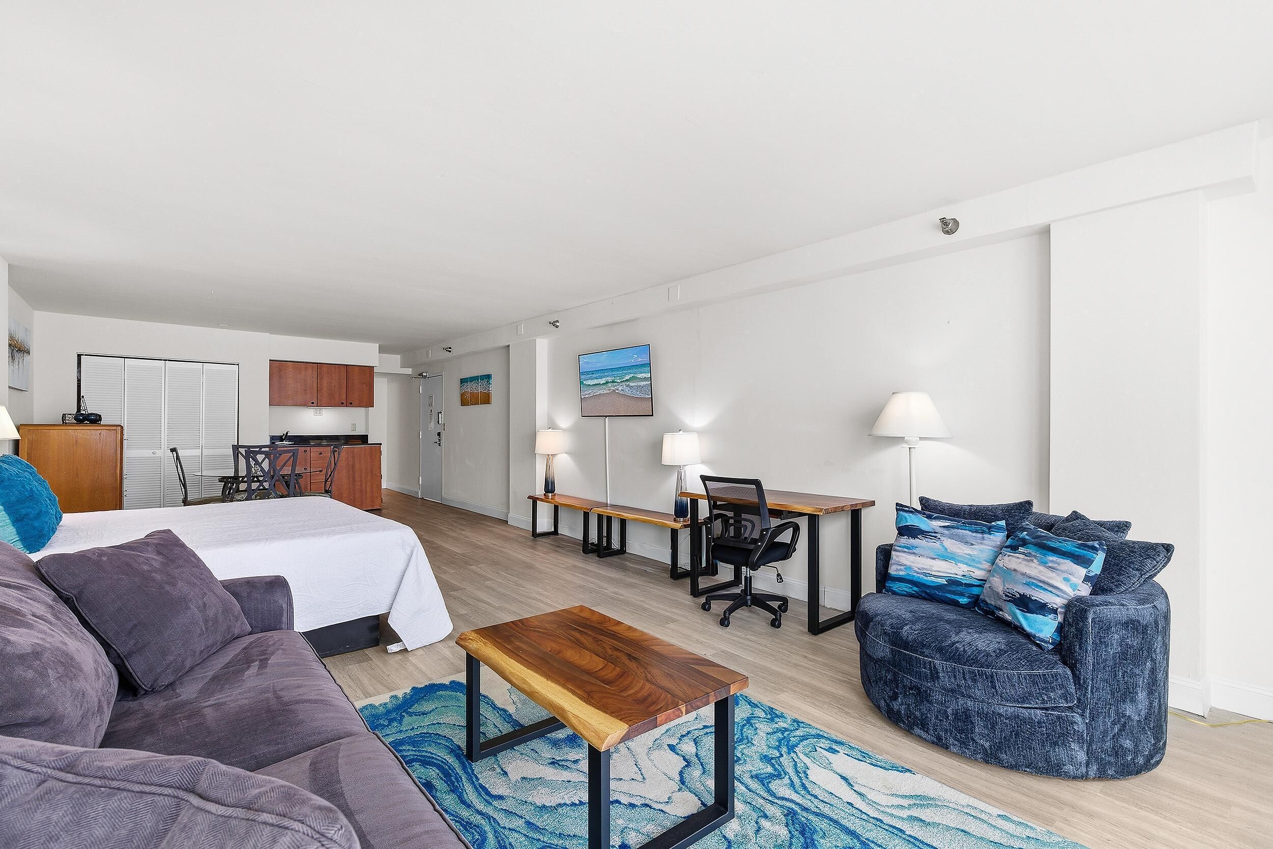 A modern hotel room with a bed, a sofa, a desk, and a chair. There is a kitchen area in the background.