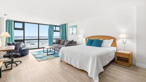 A hotel room with a large bed, a desk, a chair, a sofa, and a view of the ocean. - Oceanside Inn 313 (Daytona Beach Shores)