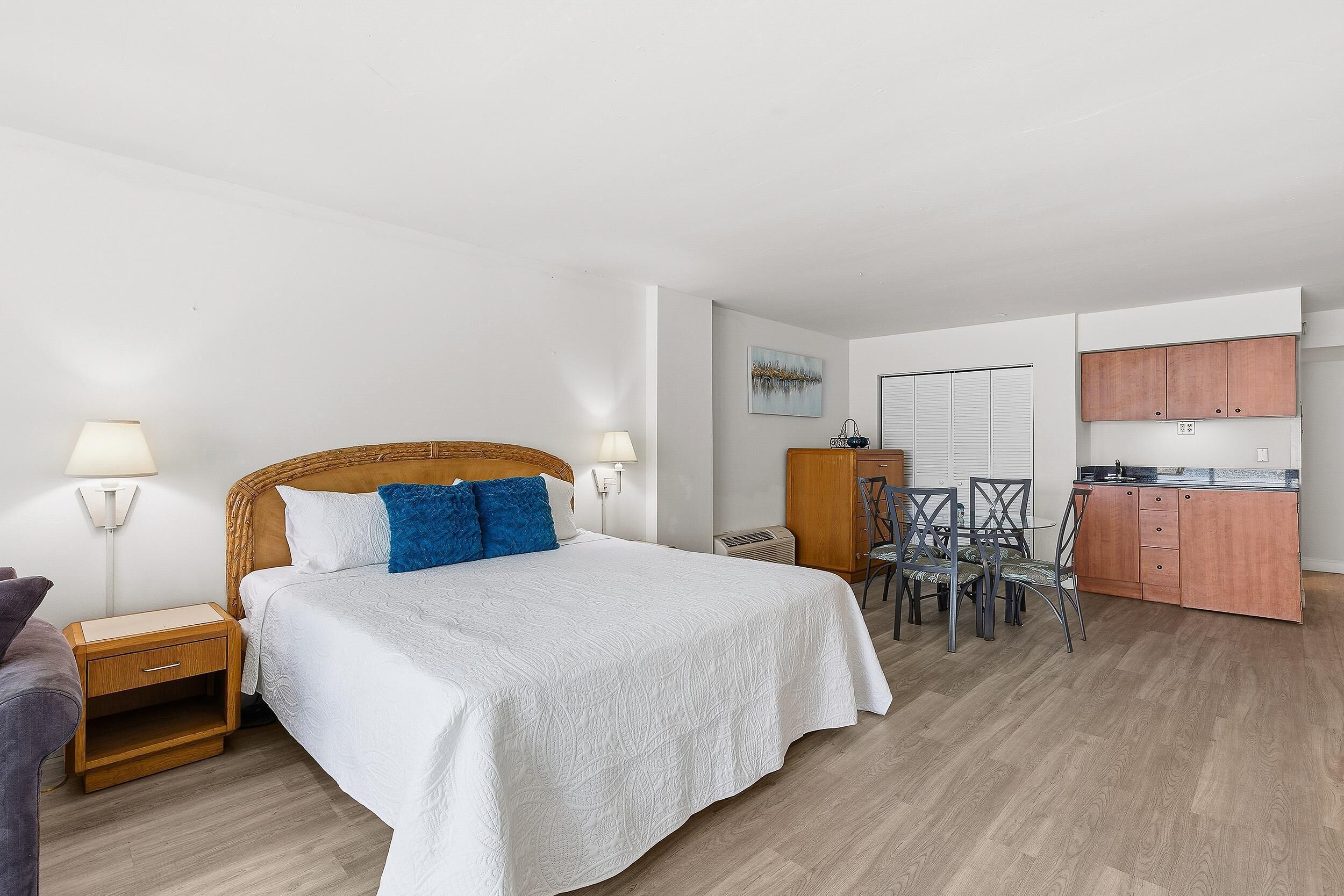 A bedroom with a bed, bedside tables, a chair, a small table, and a kitchenette area with cabinets and a countertop.