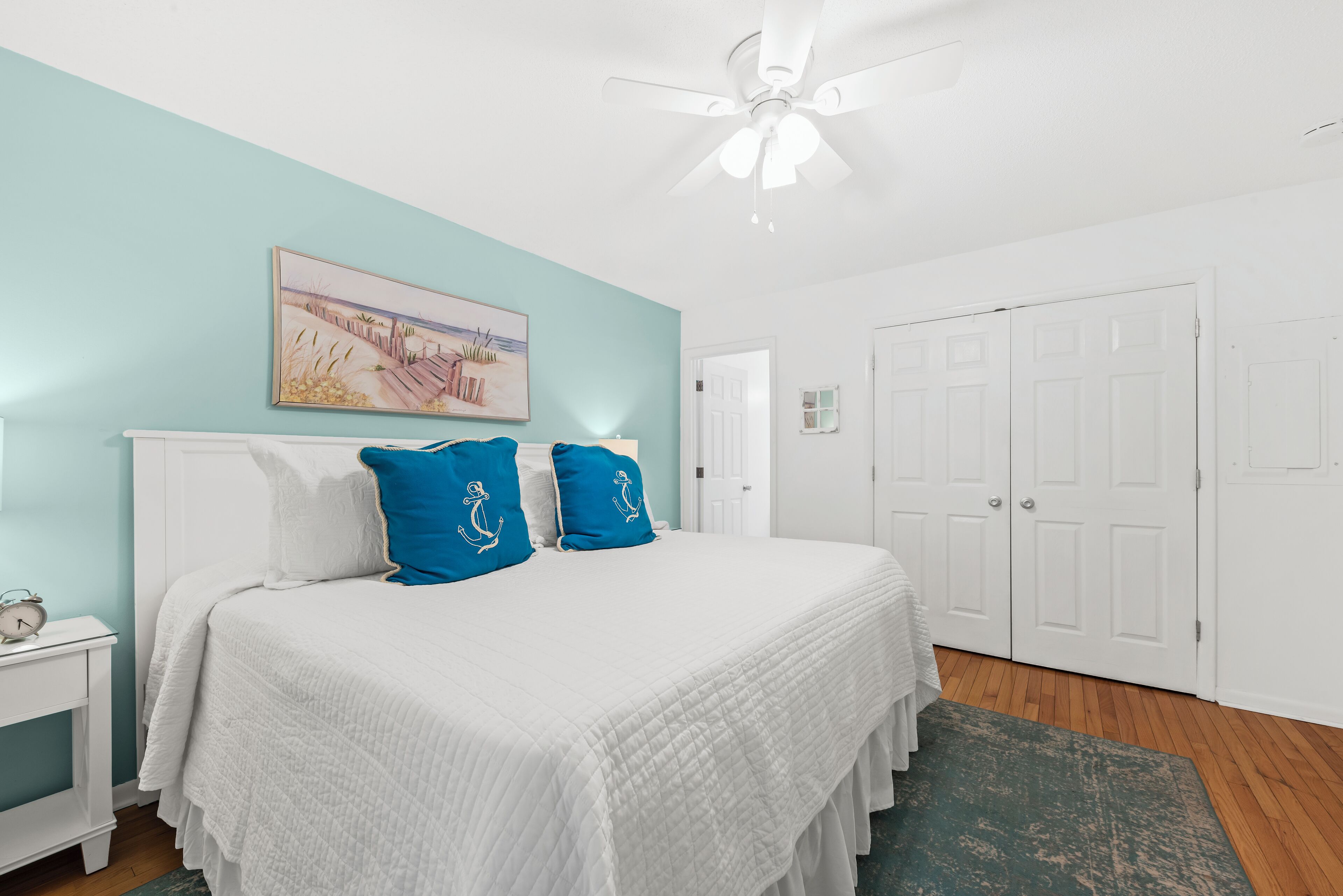 A bedroom with a bed, white nightstands, a ceiling fan, a painting, and white doors.