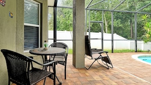 Un patio al aire libre con una mesa redonda y sillas, una piscina y vistas a árboles y una cerca blanca.