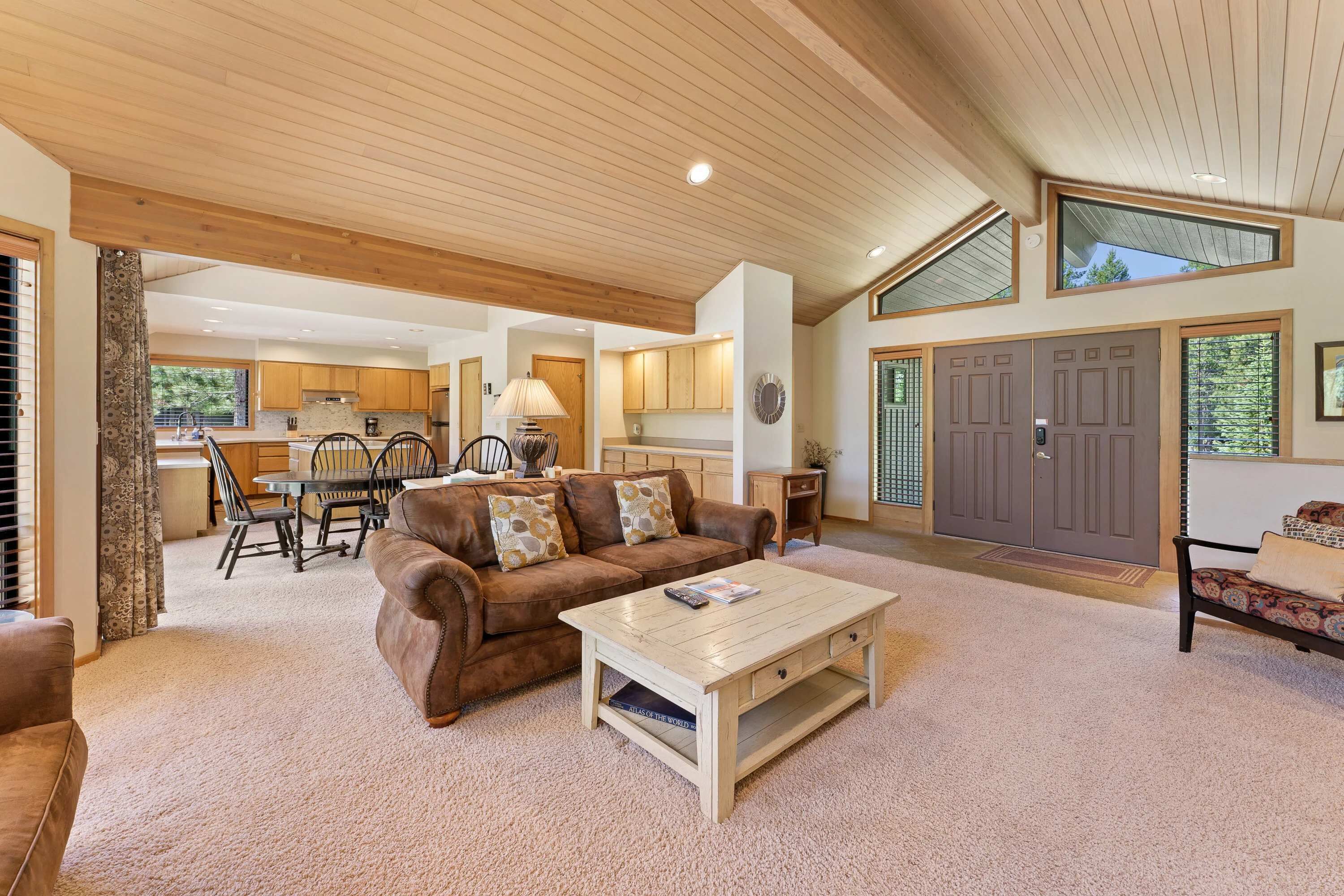 A spacious living area with a wooden ceiling, a brown leather sofa, a coffee table, and a view of the kitchen and dining area.