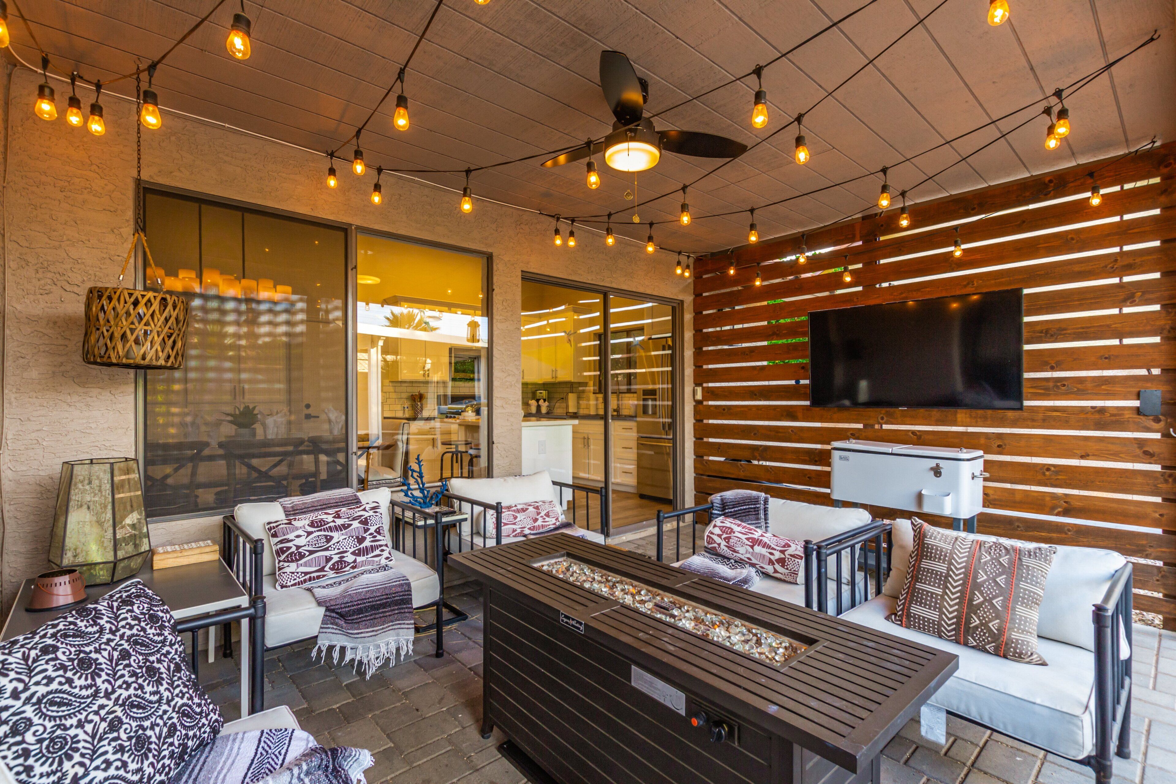 An outdoor patio area with seating, a ceiling fan, string lights, and a television mounted on the wall.