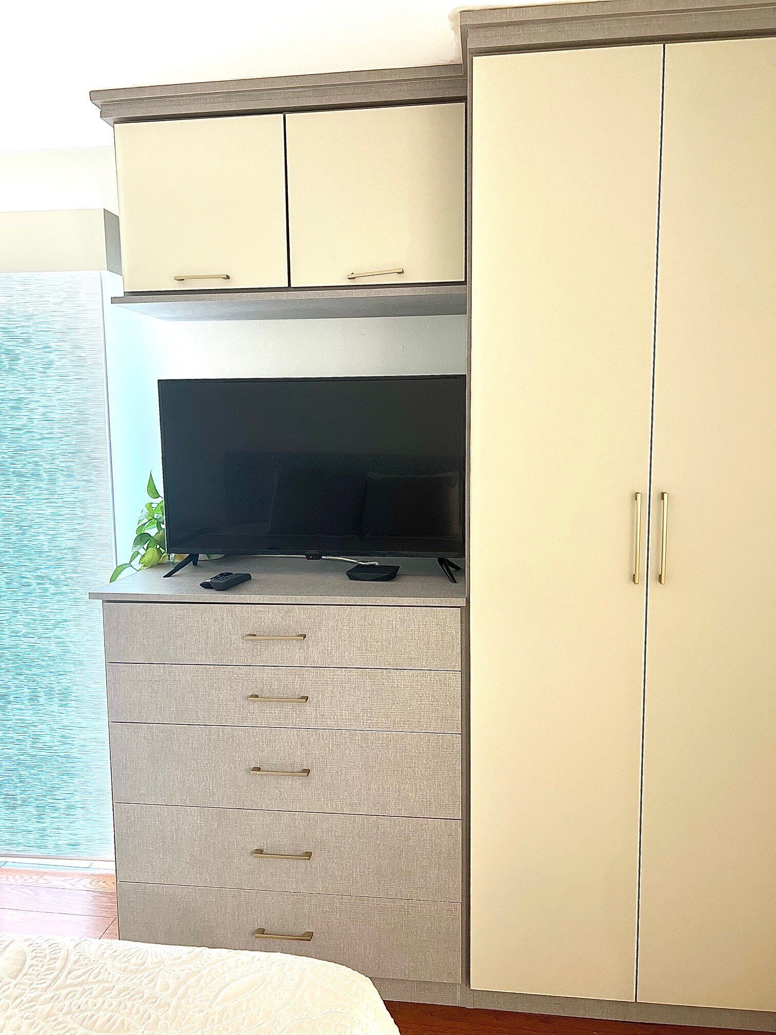 A room with a kitchenette area featuring a stove, a flat-screen TV, and a small plant. There is a large wardrobe to the right and a bed with a white bedspread.