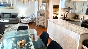 Ein modernes Wohnzimmer mit Glastisch, Sofa und einem gemusterten Sessel. Ein Essbereich mit Glastisch und Stühlen ist zu sehen. Die Küche verfügt über Granit Arbeitsplatten, eine integrierte Kochinsel und helblau lackierte Schränke.
