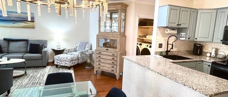 A modern living room with a glass coffee table, a sofa, and a patterned armchair. A dining area with a glass table and chairs is visible. The kitchen features granite countertops, a built-in island, and light blue cabinets.