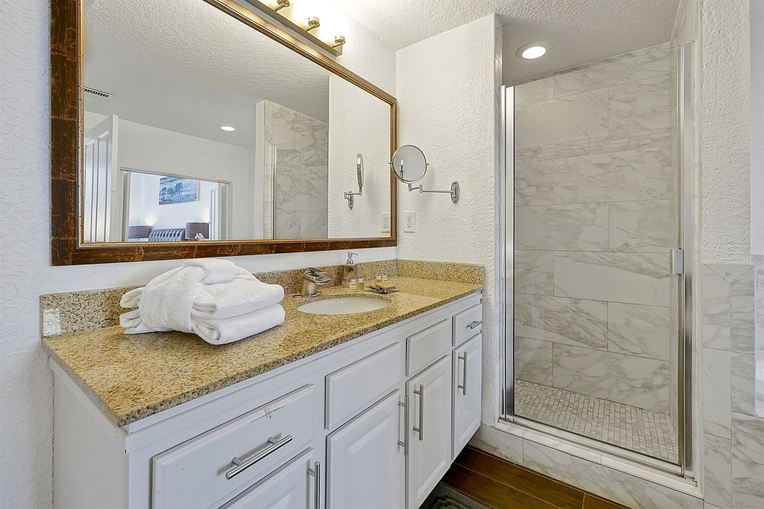 A bathroom with a granite countertop, white cabinets, a large mirror, and a glass shower enclosure.