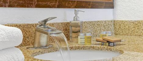 A bathroom sink with a modern faucet, a mirror above it, and a granite countertop. There are two bottles of Terra Pure shampoo and conditioner, and a wooden soap dish.