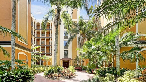 Un patio central con un sendero, rodeado de palmeras y diversas plantas, que conduce a un edificio con varios balcones.