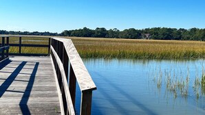 A wooden boardwalk over a calm water body with a backdrop of a dense tree line and a distant house. - 126 High Hammock (Seabrook Island)