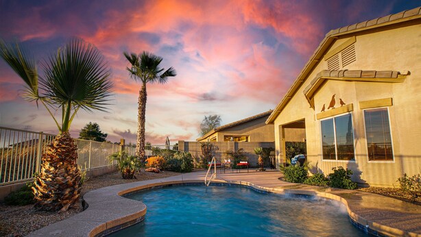 A swimming pool with a curved edge, palm trees, and a house with a large window and a bird on the roof. - 120th Avondale (Avondale)
