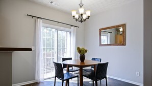 A dining area with a round table, four chairs, a vase with flowers, a chandelier, a large window with curtains, and a mirror on the wall.
