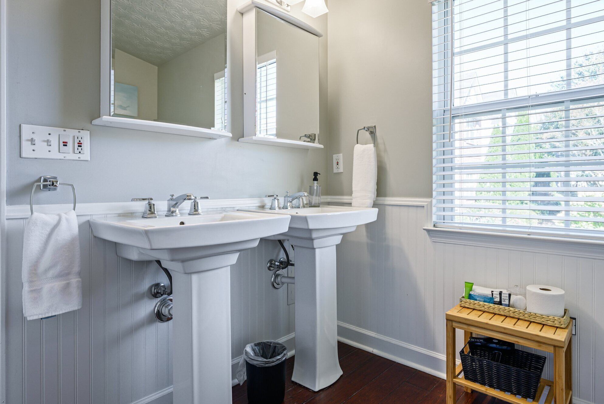 A bathroom with two sinks, a mirror, a window with blinds, a towel rack, and a small table with toiletries.