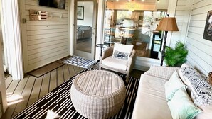 Ένας άνετος χώρος καθιστικού με έναν καναπέ από μπαμπού, ένα στρογγυλό πουφ, ένα ασπρόμαυρο ριγέ χαλί, έναν ανεμιστήρα οροφής και θέα στην κουζίνα μέσω γυάλινων θυρών.