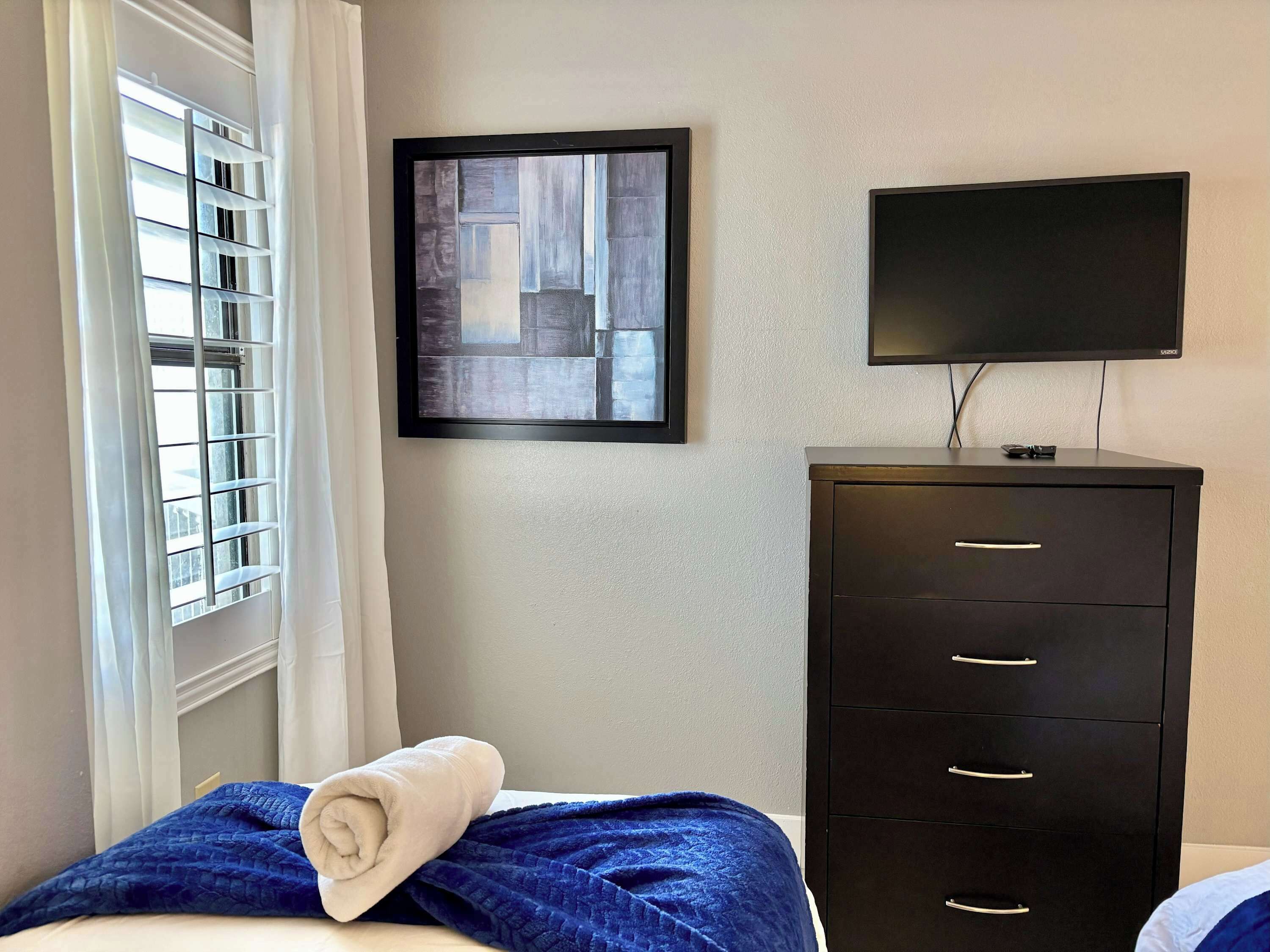 Ein Hotelzimmer mit einem Bett, einer schwarzen Kommode mit Schubladen, einem flachbildfernseher an der Wand und einem Fenster mit Jalousien.