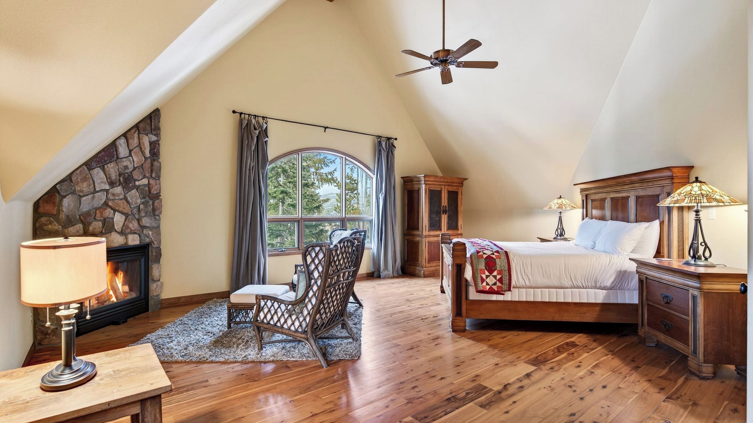 A bedroom with a stone fireplace, a bed, a nightstand, a chair, and a ceiling fan.