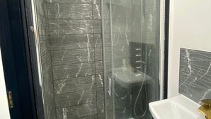 Un baño moderno con ducha envuelta en vidrio, lavamanos blanco con detalles dorados y paredes con diseño de mármol.