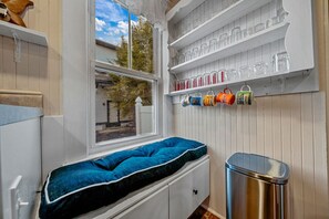 A small room with a window, a blue cushioned bench, a metal trash bin, and shelves with glassware and mugs. - Park Place in Old Orchard Beach (Old Orchard Beach)
