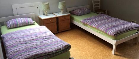Quarto com duas camas de solteiro, mesa de cabeceira com luminária, cadeira e janela com vista.