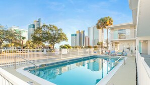 A swimming pool with a white deck, surrounded by a white fence, and a cityscape in the background. - Walk to Beach & Boardwalk • Pool • Family Suite Sleeps 8 • Full Kitchen (Myrtle Beach)