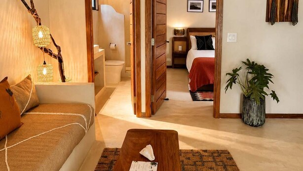 Un salón acogedor con un sofá, una mesa de centro de madera y una alfombra estampada. Hay una puerta que conduce a un dormitorio con una cama y una mesita de noche.
