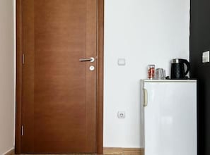Een gesloten houten deur met een handvat, een witte koelkast met een zwarte koffiezetapparaat en een glas erop, en een stopcontact aan de muur.