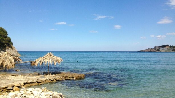 A beach with clear water, a thatched umbrella, rocky shore, and distant buildings. - Casa Mila Apartments - Studio apartment with a sea view (Ulcinj)
