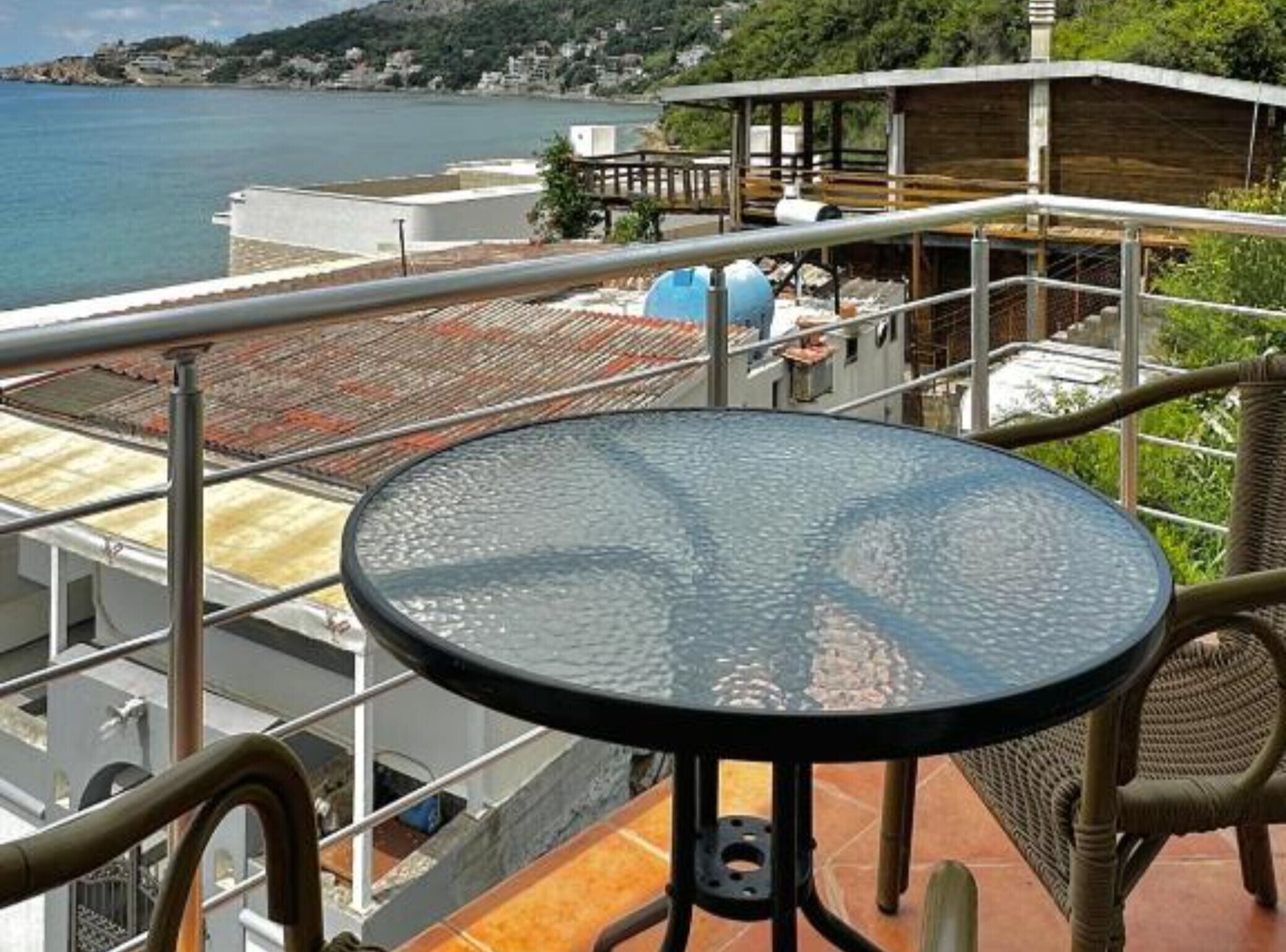 A round glass table with metal legs on a balcony overlooking a coastal area with buildings and a body of water.