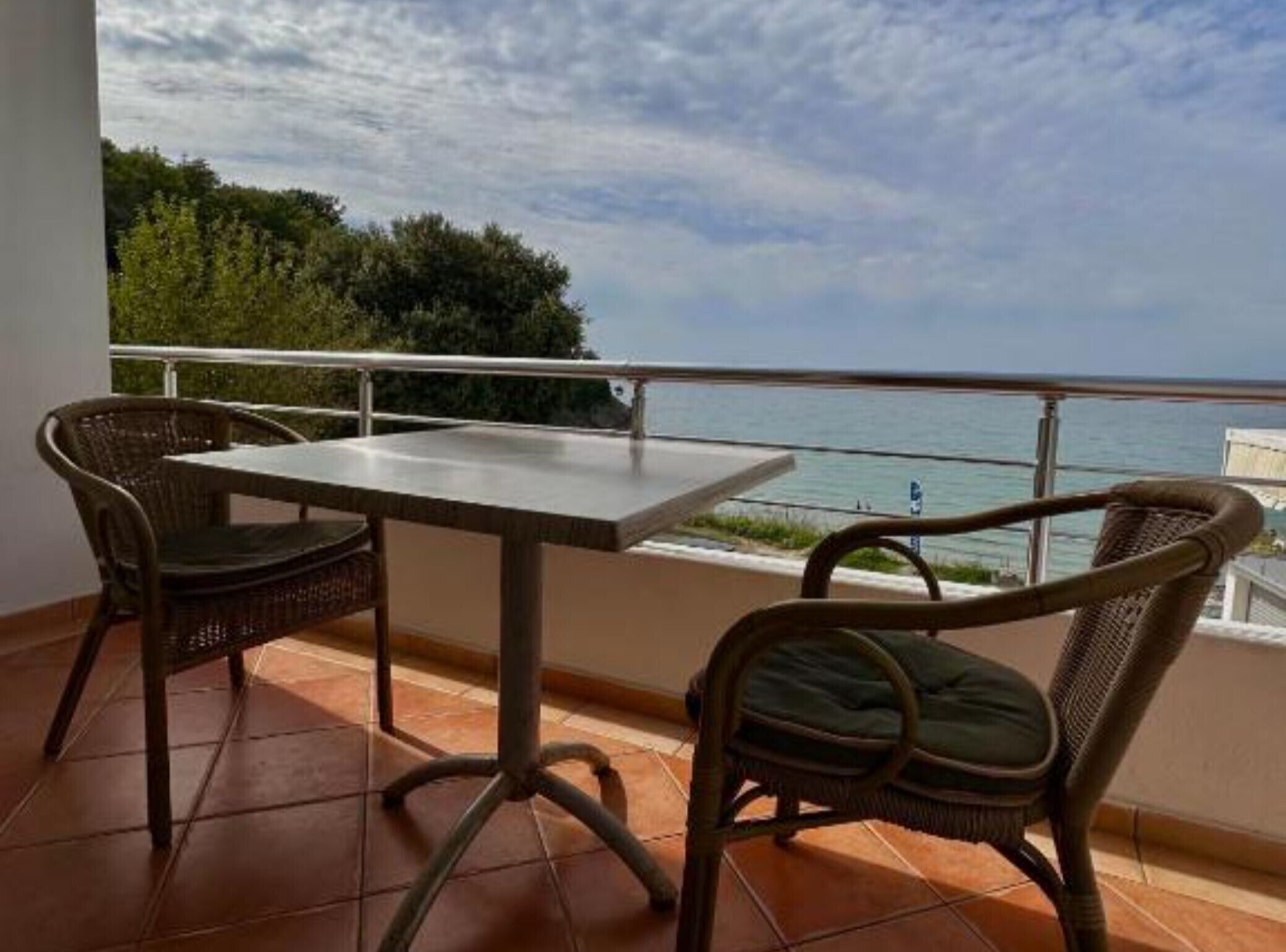 A balcony with a table and two wicker chairs, overlooking a coastal view.