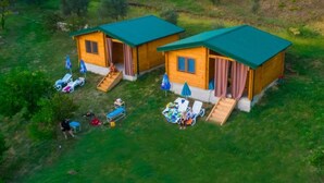 Dos cabañas de madera con tejados verdes, cada una con un pequeño porche y una sola ventana. Hay sillones de exterior y sombrillas.