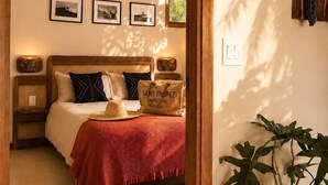 Ein Schlafzimmer mit einem Bett, einem Nachttisch, einem an der Wand befestigten Regal mit einer Flasche, gerahmten Bildern und einer Topfpflanze.
