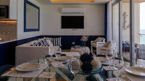 Un salon moderne avec une table à manger en verre pour deux personnes, un téléviseur à écran plat fixé au mur et un coin salon confortable avec un canapé et des fauteuils.