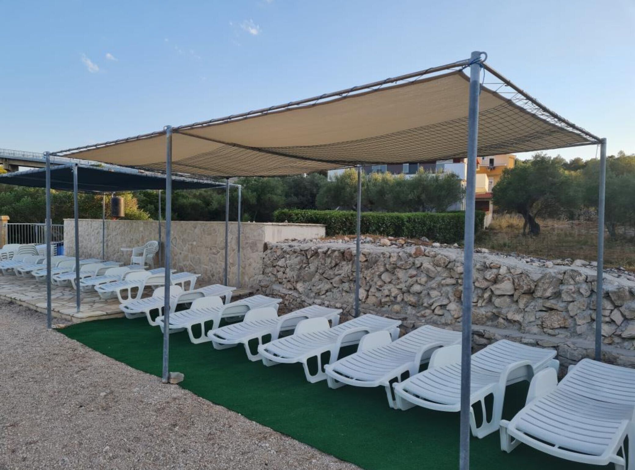 Un área de estar al aire libre con techo, sillas reclinables de plástico blanco dispuestas en filas, una pared de piedra y cielo despejado.