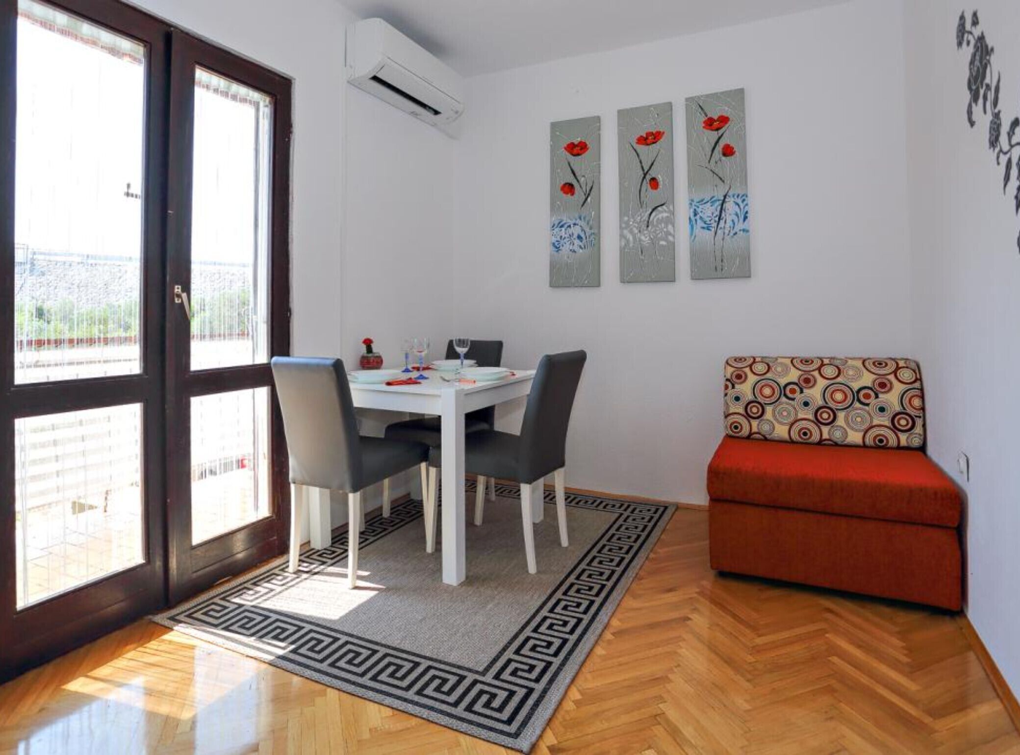 Un coin repas avec une table et des chaises blanches, un tapis à motifs et un canapé agrémenté d’un coussin rond.