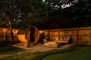 A wooden barrel sauna, a fire pit, and a sofa set in a backyard with a wooden fence. - Elmstead Retreat - Hot Tub, Game room & Kids' Play Area (Cleveland)