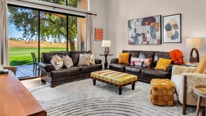 A spacious living room with a large window, a brown leather sofa, a wooden coffee table, and a white rug. - New! Beautiful Mountain-View in PGA West – 1-month Minimum Breathtaking home you’ll love to be in. (La Quinta)