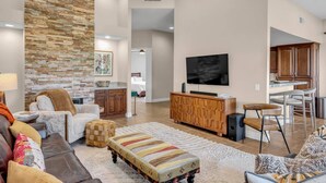 A spacious living room with a stone accent wall, a large flat-screen TV, a cozy seating area with a sofa and ottoman, and a kitchen area with wooden cabinets and a bar stool. - New! Beautiful Mountain-View in PGA West – 1-month Minimum Breathtaking home you’ll love to be in. (La Quinta)