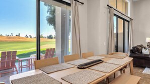 A dining area with a view of a golf course, a large window with curtains, and a leather sofa. - New! Beautiful Mountain-View in PGA West – 1-month Minimum Breathtaking home you’ll love to be in. (La Quinta)