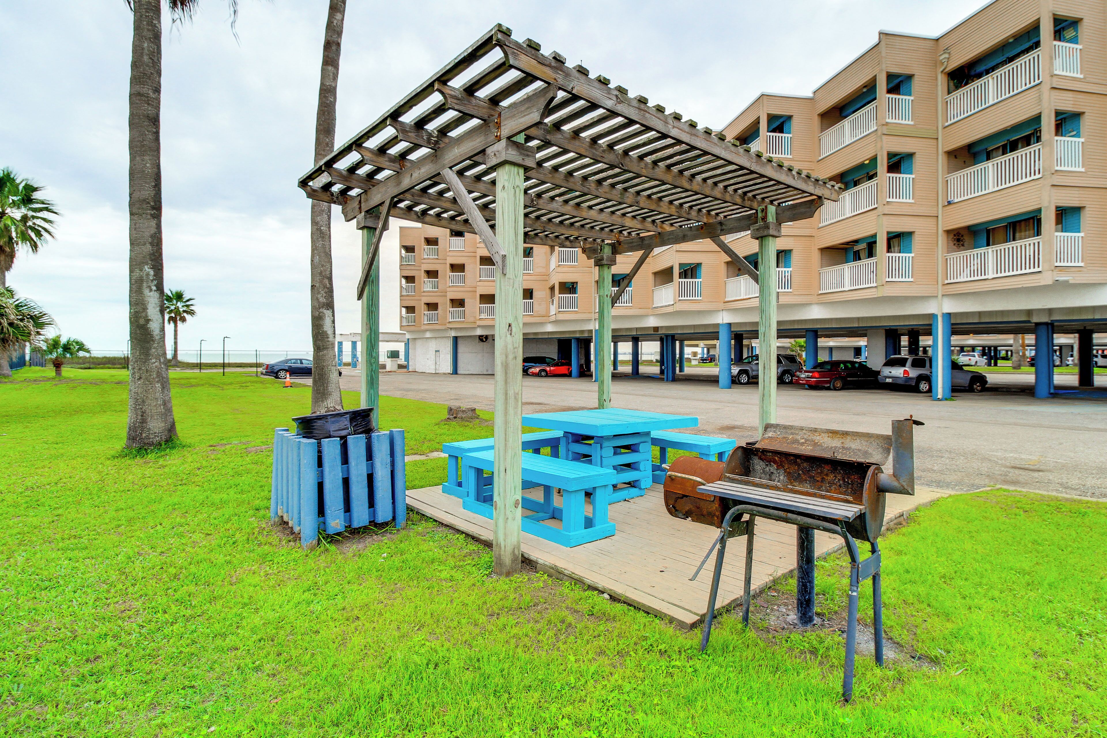 Un espace de pique-nique abrité, doté de bancs bleus et d’un barbecue, situé devant un immeuble à plusieurs étages avec des balcons.